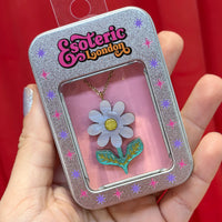 Sky blue daisy pendant in its silver gift tin with window, held in a hand against red fabric