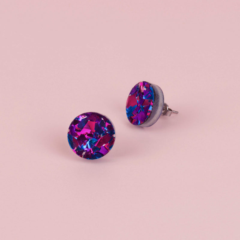 Pair of round purple confetti glitter stud earrings with pink and blue flecks, on pale pink paper