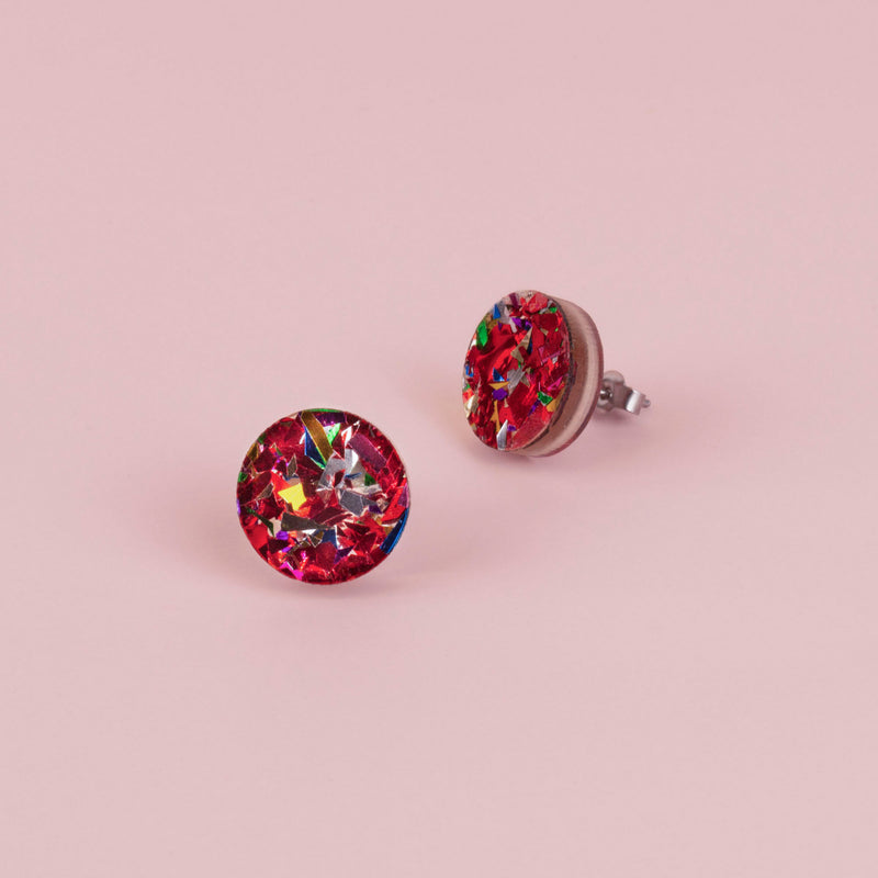Pair of round red confetti glitter stud earrings with silver, gold and green flecks, on pale pink paper
