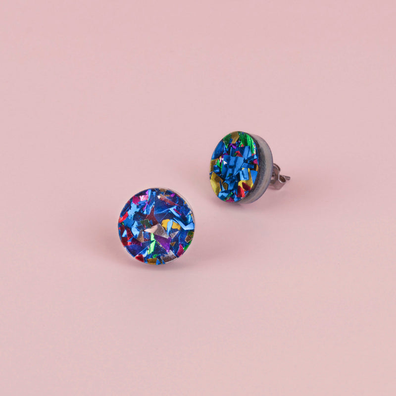 Pair of round blue confetti glitter stud earrings with silver, green and pink flecks, on pale pink paper