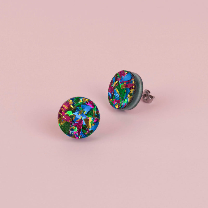Pair of round green confetti glitter stud earrings with blue, pink and gold flecks, on pale pink paper
