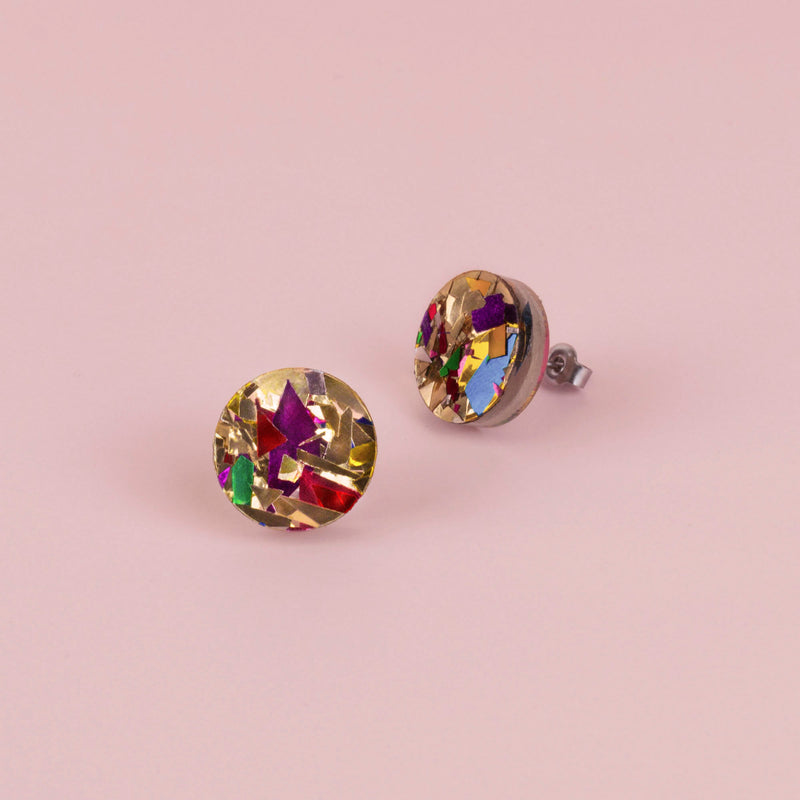 Pair of round gold confetti glitter stud earrings with red, purple and green flecks, on pale pink paper