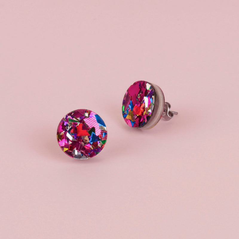 Pair of round pink confetti glitter stud earrings with blue, silver and red flecks, on pale pink paper