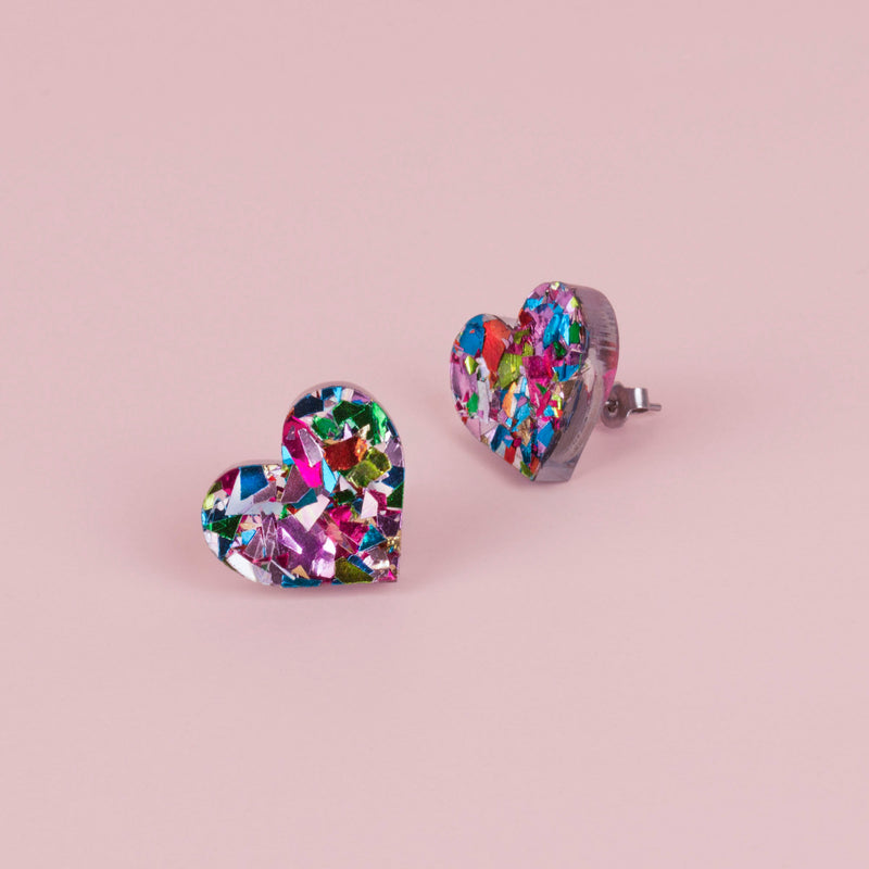 Pair of rainbow confetti glitter heart stud earrings with pink, green, blue and lilac flecks, on pale pink paper