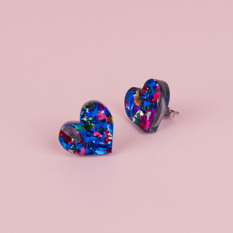Pair of blue confetti glitter heart stud earrings with pink, silver and green flecks, on pale pink paper