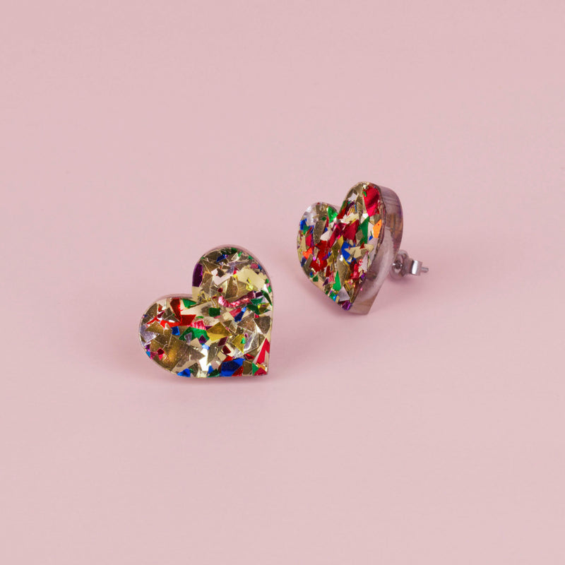 Pair of gold confetti glitter heart stud earrings with red, green and blue flecks, on pale pink paper