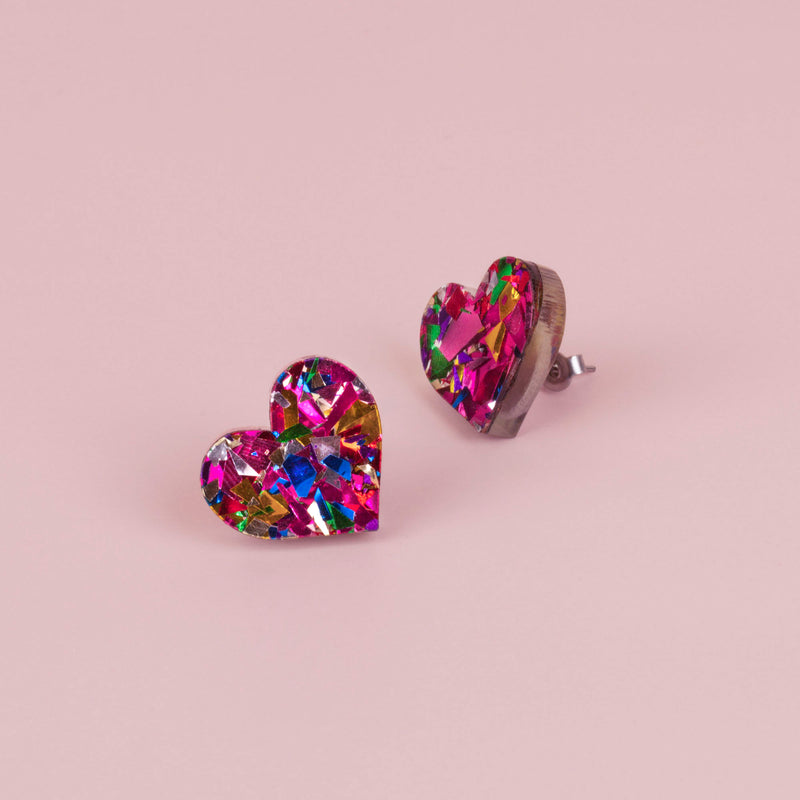 Pair of pink confetti glitter heart stud earrings with blue, gold and silver flecks, on pale pink paper