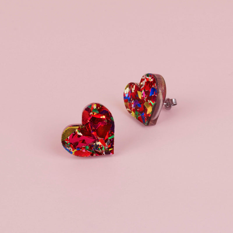 Pair of red confetti glitter heart stud earrings with gold, green and blue flecks, on pale pink paper