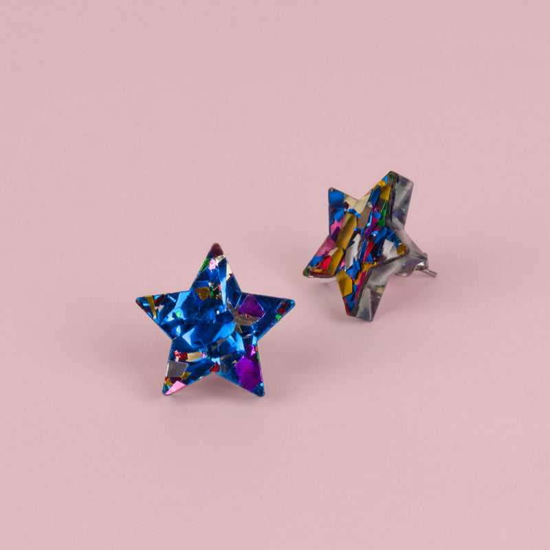 Pair of blue confetti glitter star stud earrings with purple, gold and pink flecks, on pale pink paper