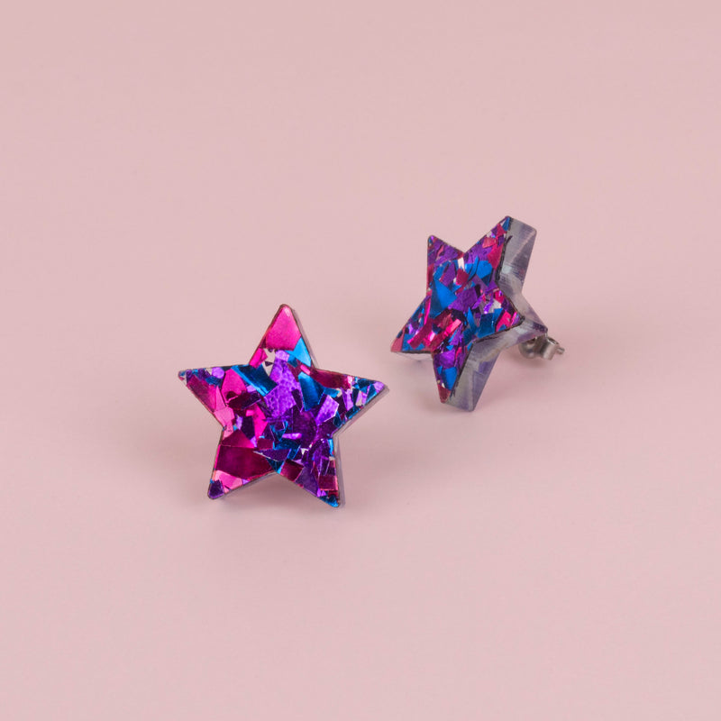 Pair of purple confetti glitter star stud earrings with pink and blue flecks, on pale pink paper