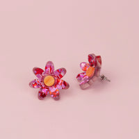Pair of pink and red holographic confetti flower studs with orange wood effect centres on pink paper, one on its side