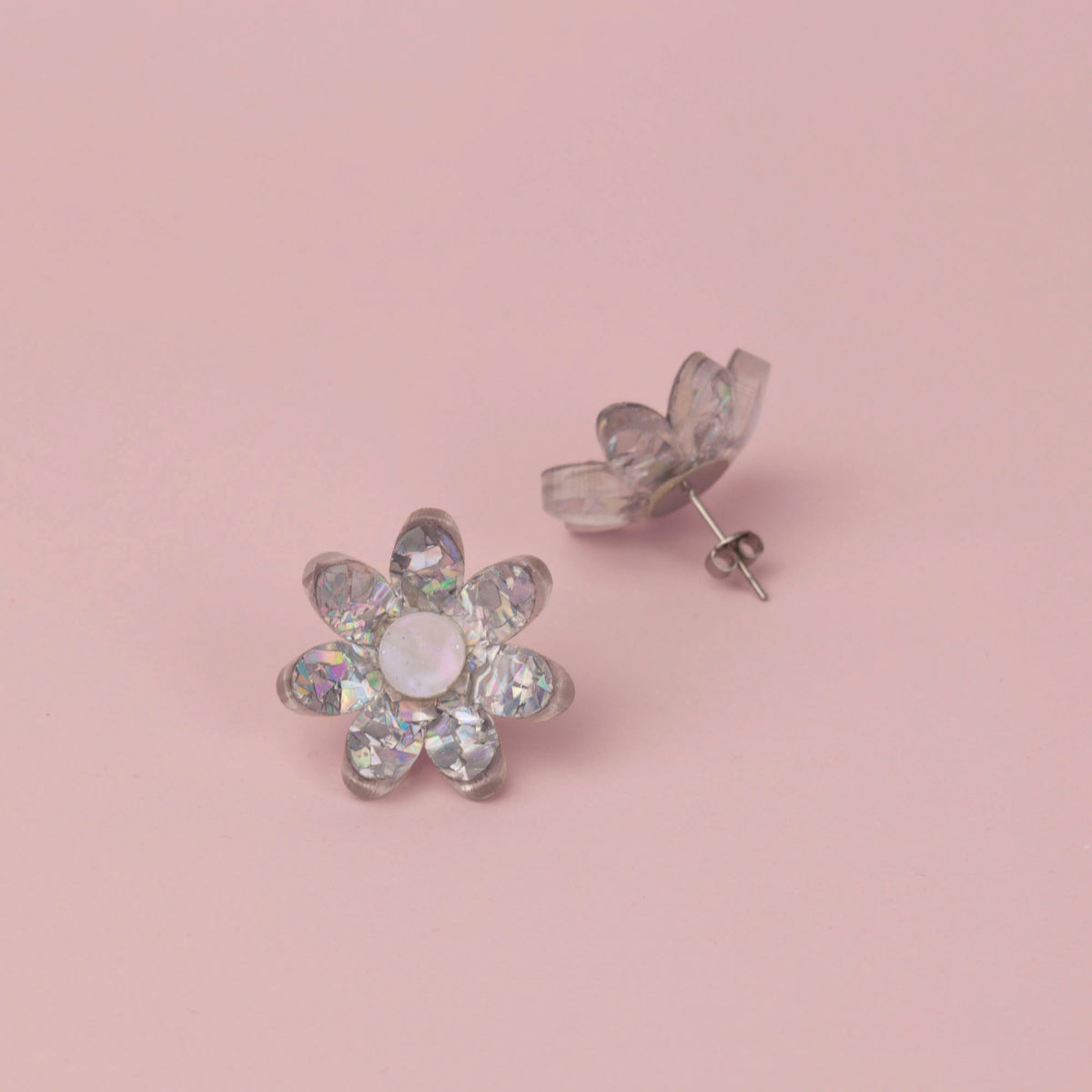 Silver holographic flower stud face up beside its pair showing the post and butterfly back, on pink paper