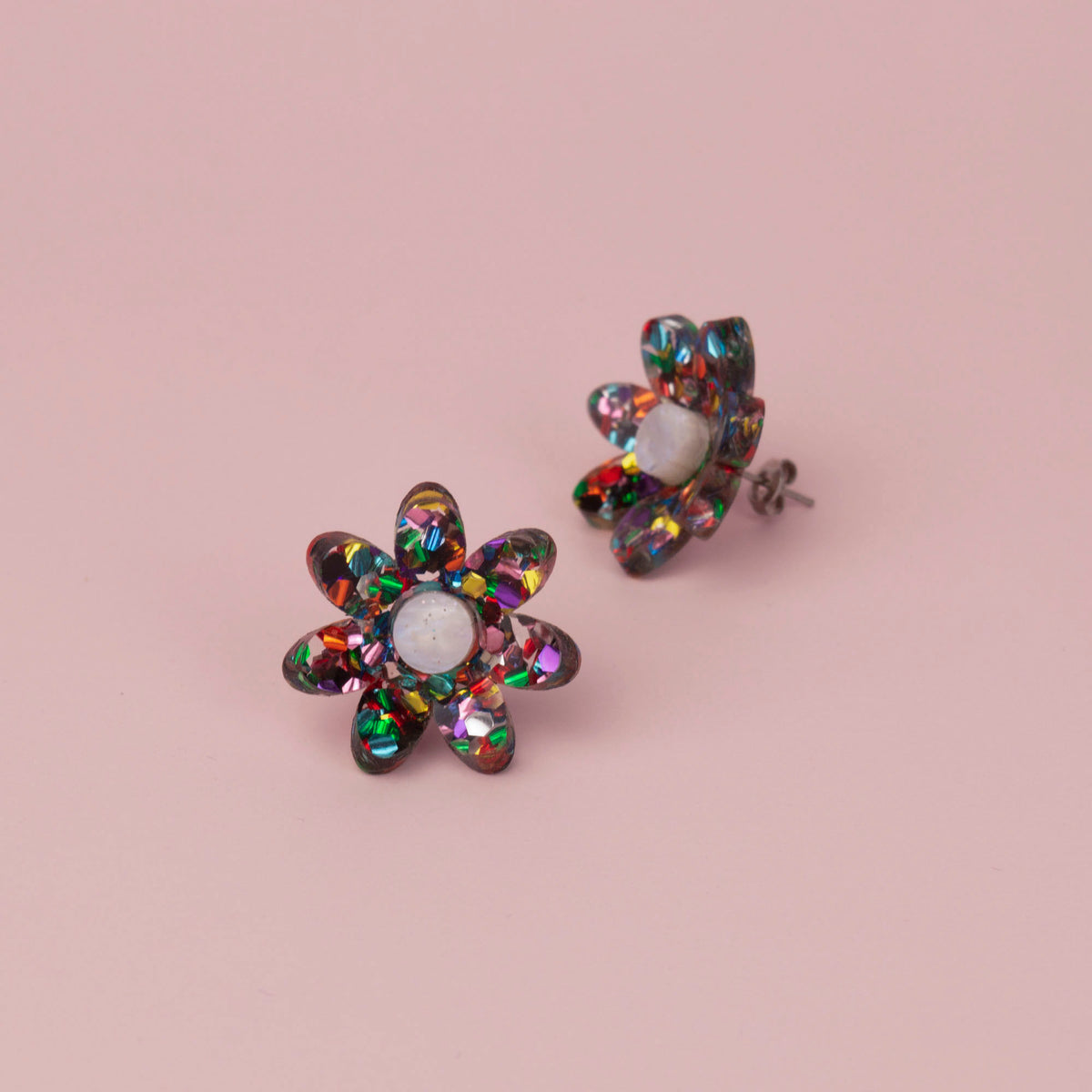 Rainbow confetti flower studs with multicoloured glitter petals and white centres on pink, one tilted to show the post