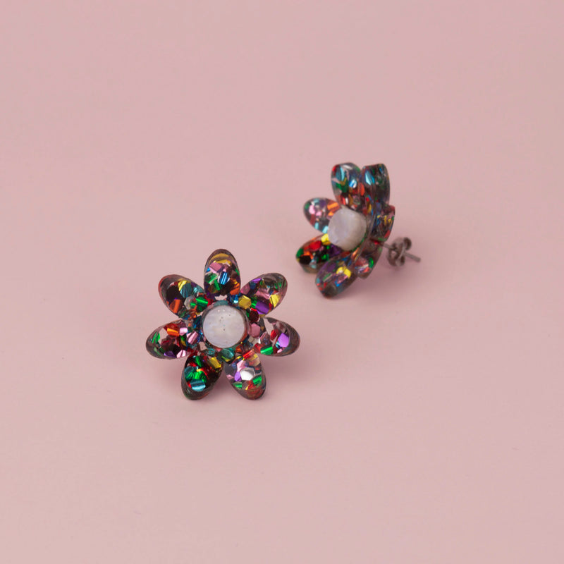 Rainbow confetti flower studs with multicoloured glitter petals and white centres on pink, one tilted to show the post