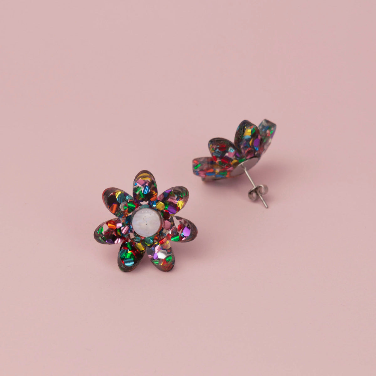 Rainbow confetti flower stud face up beside its pair standing on its post, on pink paper