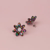 Rainbow confetti flower stud face up beside its pair standing on its post, on pink paper