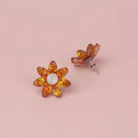 Orange holographic confetti flower stud lying face up beside the second earring showing its silver post, on pink