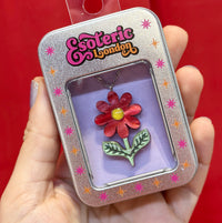 Scarlet red daisy pendant in its silver gift tin with window and lilac backing, held in a hand against red