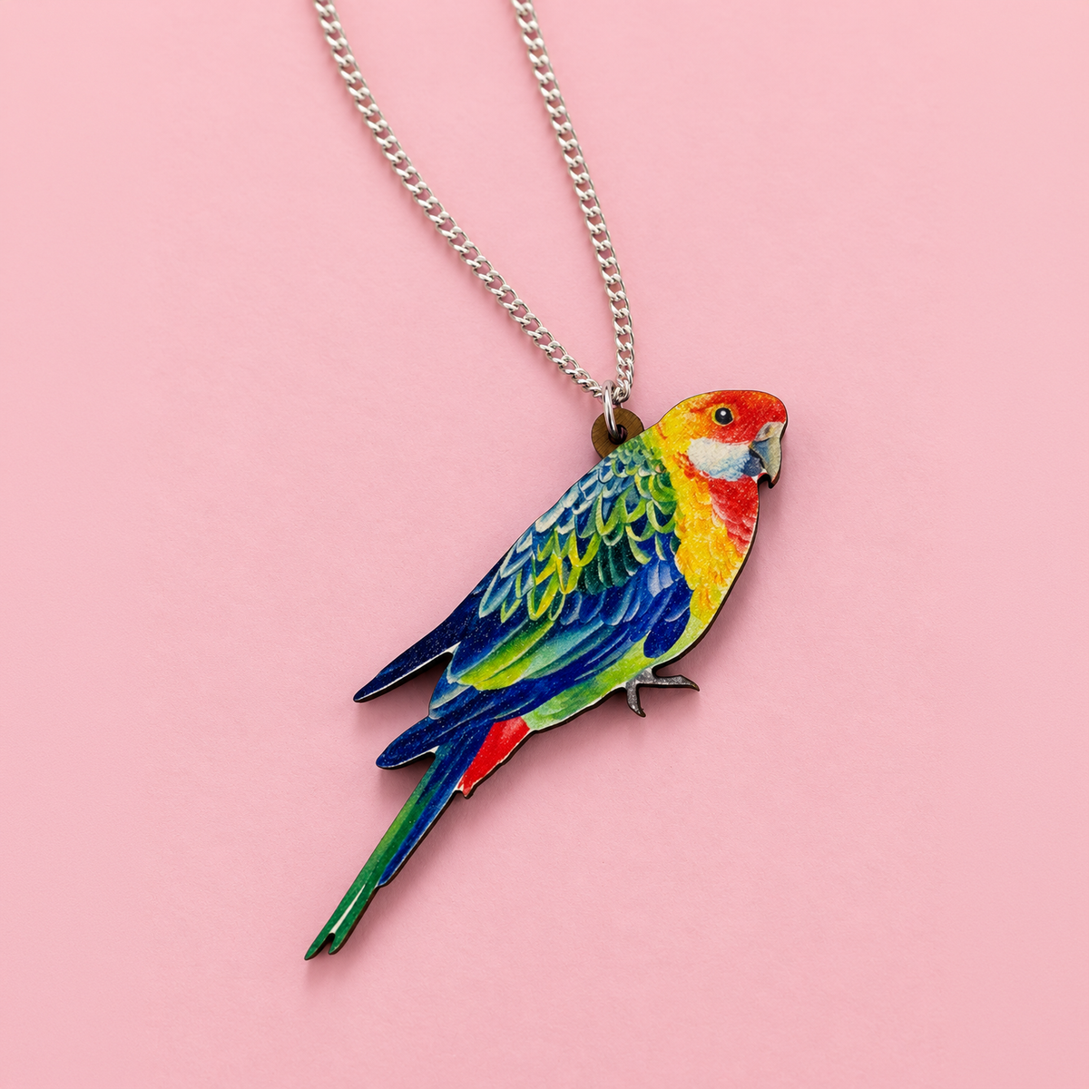 Rosella parrot pendant with red head, yellow chest and blue-green wings, on a silver curb chain over pink paper