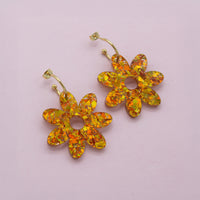 Orange and gold holographic glitter flower dangle earrings with gold hoop posts, flat lay on pale lilac paper