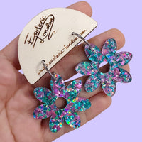 Lilac and turquoise holographic flower hoop earrings held in a hand on a wooden card, against lilac