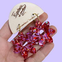 Pink and red holographic confetti flower hoop earrings held in a hand on a wooden card, against lilac