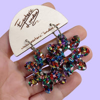 Rainbow confetti flower hoop earrings held in a hand on a wooden card, against a lilac backdrop