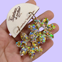 Gold and silver holographic confetti flower hoop earrings held in a hand on a wooden card, against lilac
