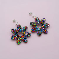 Rainbow glitter confetti flower dangle hoop earrings with silver hoops, laid flat on pale pink paper