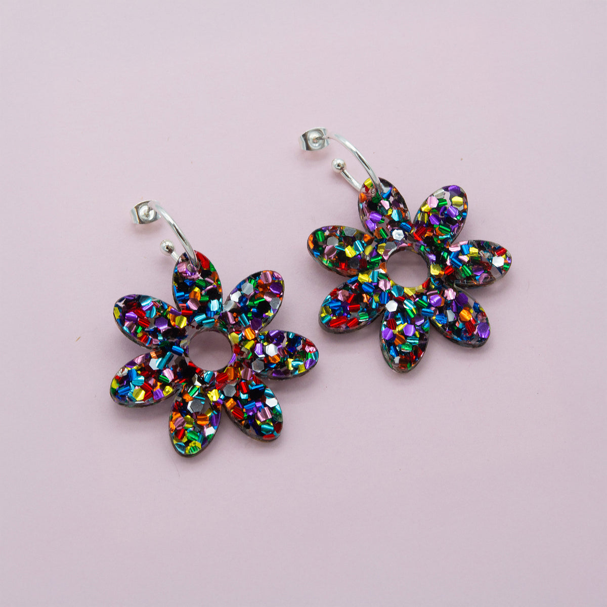 Rainbow glitter confetti flower dangle hoop earrings with silver hoops, laid flat on pale pink paper