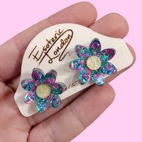 Lilac and turquoise confetti flower studs with pale yellow centres on a wooden backing card in a hand, pink backdrop