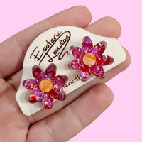 Pink and red confetti flower studs held in a hand on a wooden backing card, against pink