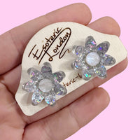 Silver holographic confetti flower studs on a wooden backing card held in a hand, against a pink backdrop
