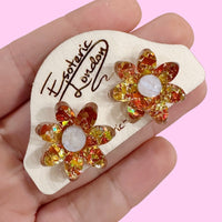 Orange holographic confetti flower studs held in a hand on a wooden backing card, against pink