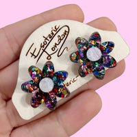 Rainbow confetti flower studs held in a hand on a wooden backing card, against pink