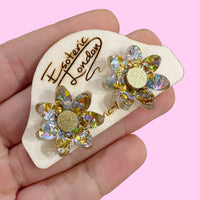 Silver and gold holographic flower studs with gold glitter centres on a wooden backing card in a hand, pink backdrop
