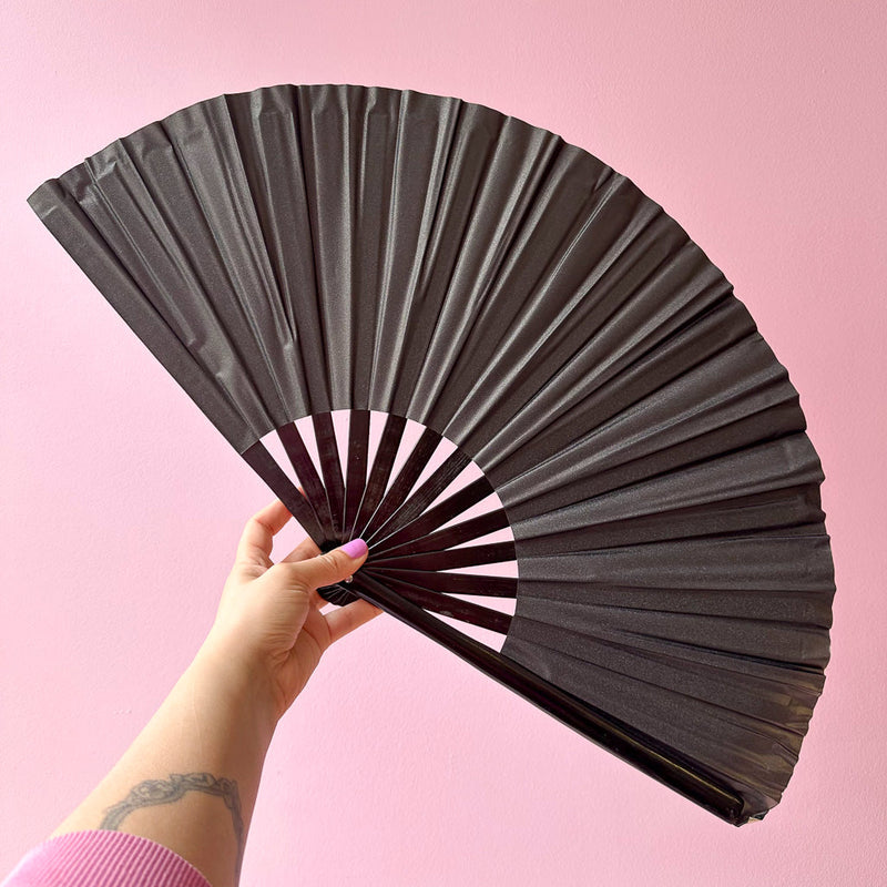 Giant black matte hand fan with black bamboo handle, held open in a hand against a pink wall