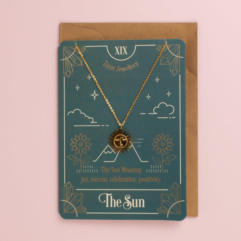 Gold sun face pendant necklace on a teal The Sun tarot card with sunflowers and mountains, kraft envelope behind