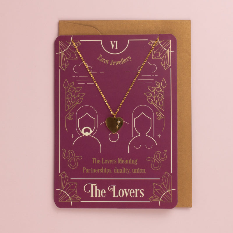 Gold heart pendant necklace on a plum The Lovers tarot card with two figures and snakes, kraft envelope behind