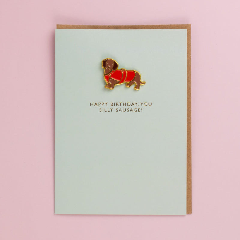 Pale green birthday card with a gold edged enamel pin of a dachshund in a red coat, kraft envelope behind