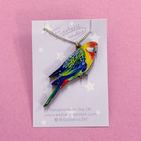 Rosella parrot pendant necklace on a lilac star printed backing card, on a pink background