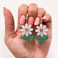 Hand holding white daisy dangle earrings with orange centres and green leaves on silver huggie hoops