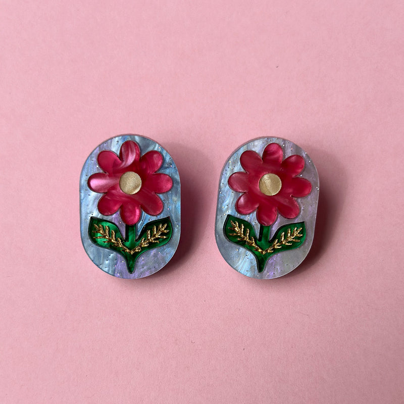 Pair of oval studs with a pink flower, gold centre and green leaves on pearly blue acrylic, flat on pink paper