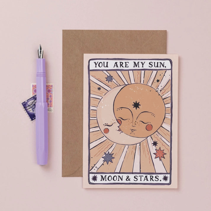 Sun, Moon and Stars tarot style card with kraft envelope on pink paper, next to a lilac fountain pen and vintage stamps