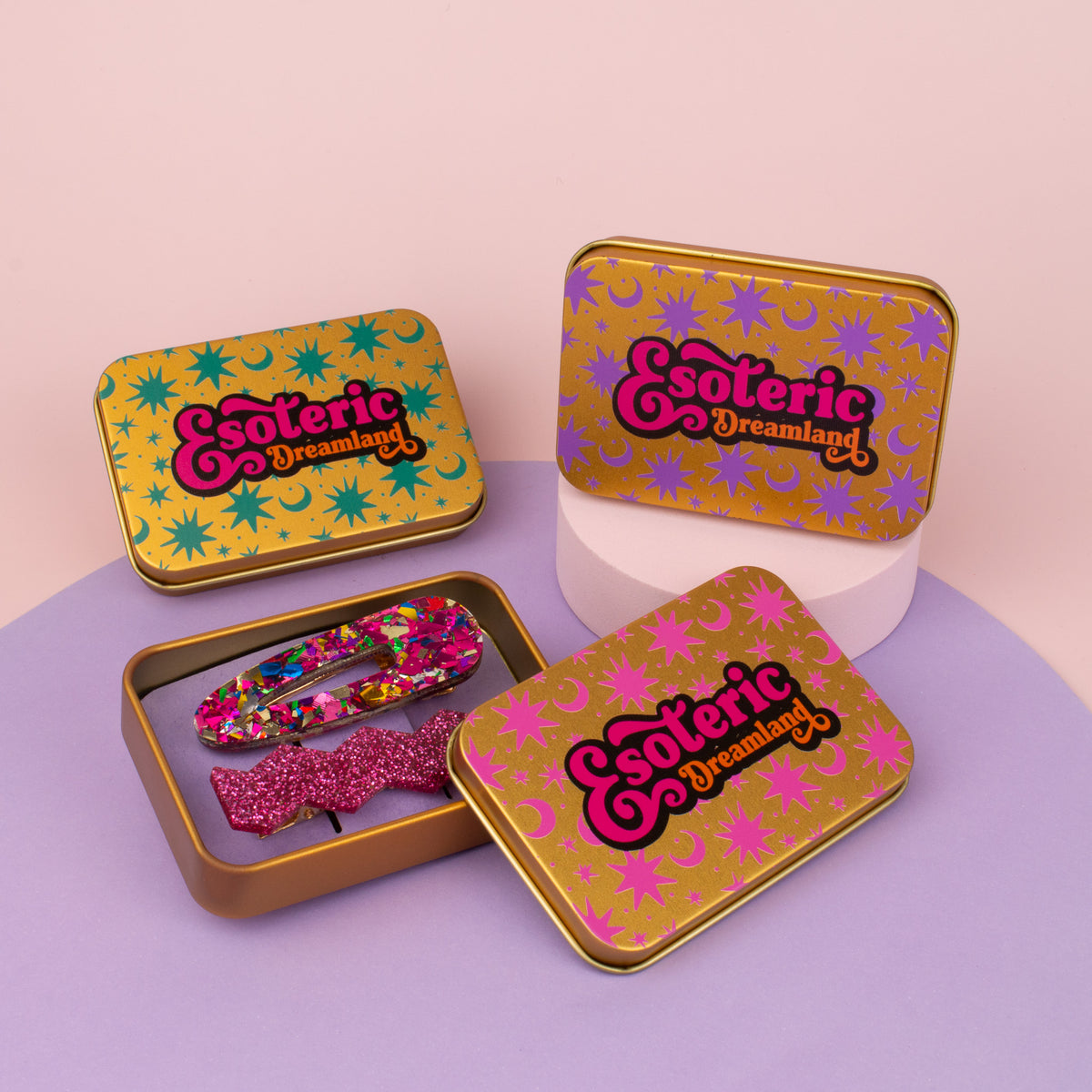 Gold tin with its lid off showing two glitter hair clips, other printed tins stacked beside it on a pink backdrop